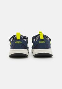 Tênis azul-marinho com detalhes em amarelo, painéis de malha, alças no calcanhar e sola branca. A visão traseira destaca os texturas e cores contrastantes.