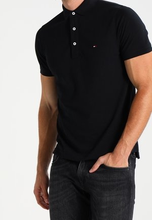 Homme portant un polo noir ajusté à trois boutons et un jean foncé, une main dans la poche, sur un fond blanc uni.