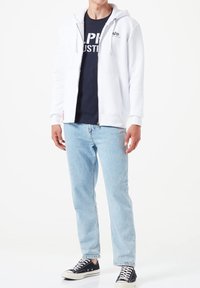 Felpa bianca con zipper e tasche frontali sopra una maglietta blu scuro. Jeans in denim azzurro e sneakers nere completano l'outfit casual.