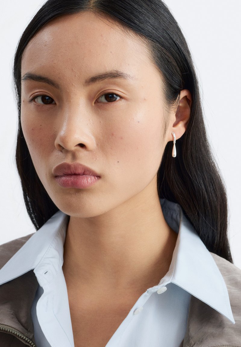Femme aux longs cheveux foncés portant des boucles d'oreilles argentées en forme de goutte, une chemise bleu clair à col et une veste marron, regardant droit devant avec une expression neutre.