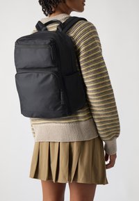 Černý nylonový batoh s několika zipovými přihrádkami a přední kapsou, nošený přes béžový pruhovaný svetr a khaki skládací sukni.