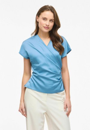 Femme aux longs cheveux bruns portant un chemisier bleu clair à manches courtes et un pantalon blanc, debout devant un fond blanc uni.