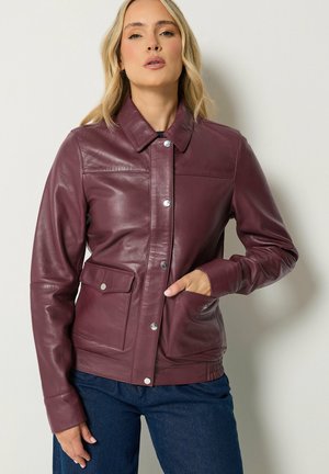 Frau trägt eine violette Lederjacke mit Druckknöpfen und Klappentaschen, kombiniert mit dunkelblauen Jeans, steht vor einer schlichten Wand.