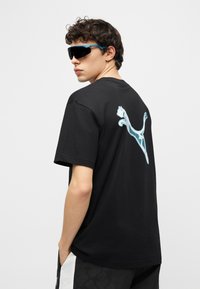 Νέος άνδρας με μαύρο t-shirt με ανοιχτό μπλε λογότυπο Puma στην πλάτη, μαύρο και άσπρο σορτς και μεγάλα μαύρα γυα λιά ηλίου, κοιτάζοντας πάνω από τον ώμο του.