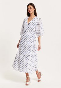 White wrap dress with blue polka dots and floral embroidery, featuring short, flutter sleeves and a tie at the waist.