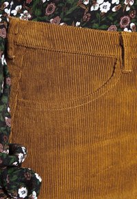 Brun kordfløjsstof med lodret ribbing, der har en sidelomme og et blomstermønster af accentstof i sort, grøn og hvid.