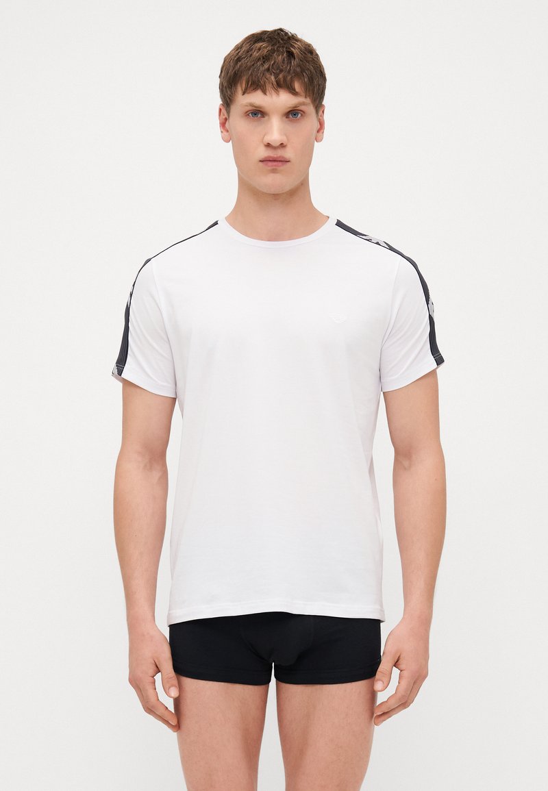 White cotton T-shirt with black side stripes, crew neckline, and short sleeves, worn with black shorts. Smooth texture, casual fit.