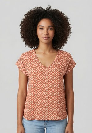 Donna con capelli ricci che indossa una maglietta a maniche corte con scollo a V e motivo arancione e beige, e jeans azzurri, in piedi davanti a uno sfondo semplice.