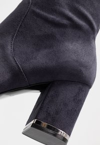 Kraftigt marinblå mocka ankelsko med en fyrkantig klack som har en metallisk accent vid basen. Slät textur och elegant silhuett.