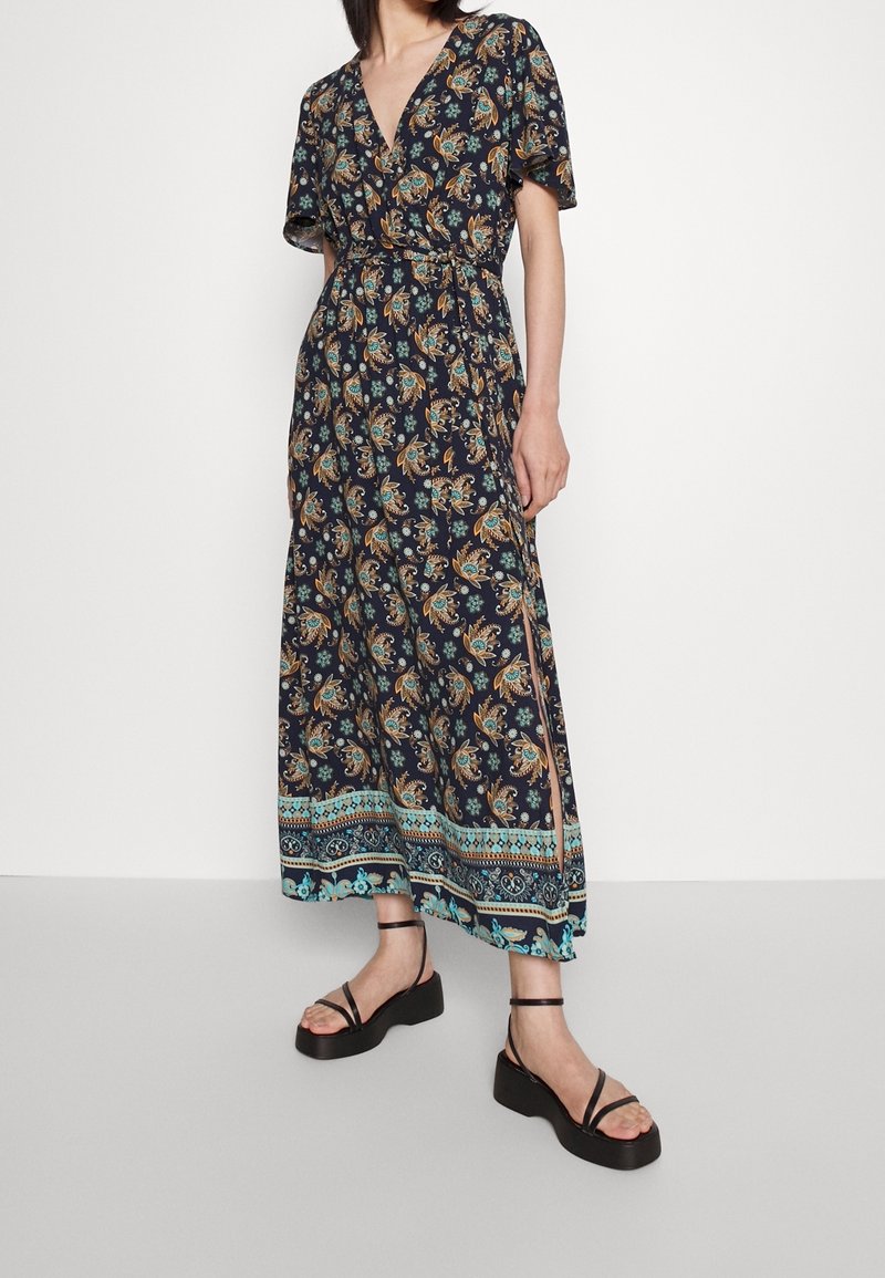 Lång mörk blommig klänning med v-ringning, korta ärmar och cinchad midja. Har blå och orange paisleymönster samt sidoklipp. Bärs med svarta sandaler.