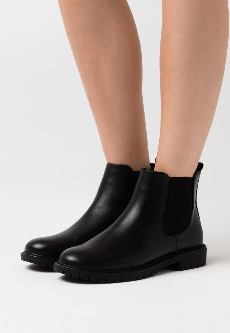Bottes Chelsea en cuir noir avec panneaux latéraux élastiques portées sur jambes nues sur un fond clair uni.