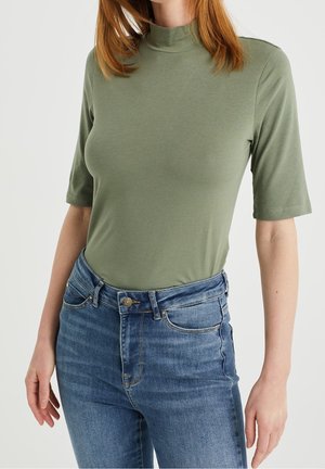 Nainen, joka on pukeutunut oliivinvihreään lyhythihaiseen korkean kaula-aukon paitaan, joka on taitettu korkeavyötäröisten sinisten farkkujen sisään, yksinkertaista taustaa vasten.