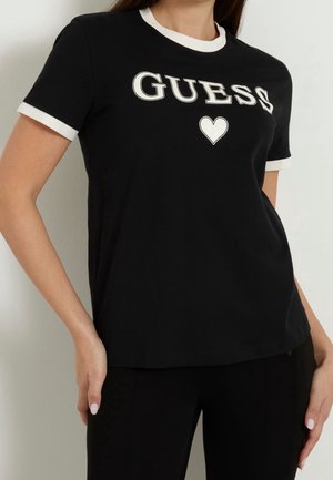Kvinde iført en sort T-shirt med korte ærmer og hvide kraver og manchetter, med teksten "GUESS" og et hvidt hjerte på brystet.