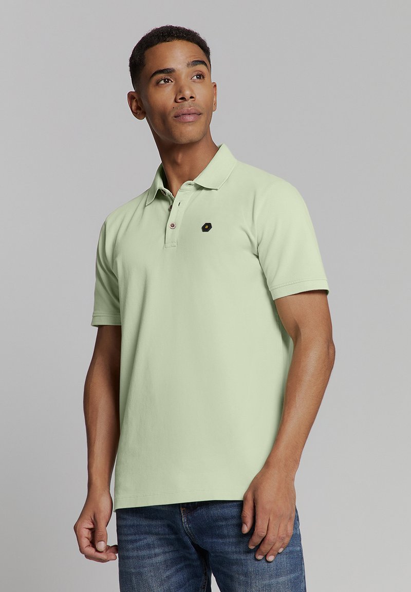 Lichtgroene polo met een klassieke kraag, twee knopen en een klein zwart logo-patch op de borst; gecombineerd met een blauwe jeans.