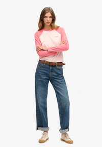 Jongevrouw met lang haar staat met gekruiste armen, draagt een roze-wit shirt met lange mouwen, blauw opgerolde spijkerbroek, een bruine riem en beige sneakers.