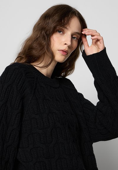 Jeune femme aux longs cheveux bruns portant un pull en maille torsadée noir, touchant sa tempe avec sa main droite sur un fond uni.