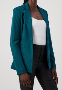 Mörkhyad person som bär en teal kavaj över en vit ribbad tanktop och svarta åtsittande byxor, stående mot en enfärgad bakgrund.