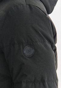 Czarna kurtka puchowa z teksturowaną powierzchnią, charakterystyczną okrągłą czarną naszywką z logo na rękawie oraz elastycznym dołem, który zapewnia dopasowane wykończenie.