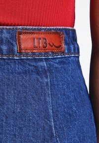 Džínové kalhoty s vysokým pasem, modré barvy a texturovanou tkaninou. Na zadní straně mají hnědou koženou nášivku s nápisem "LTB".