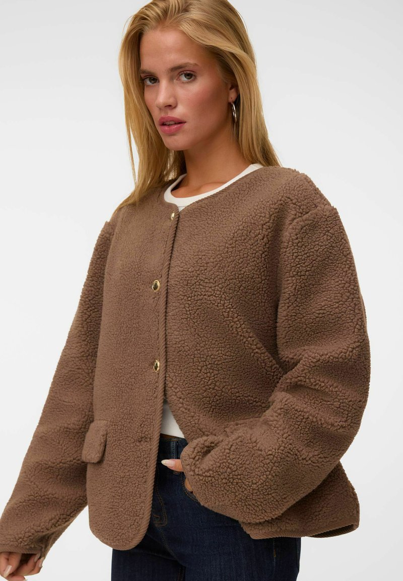 Jeune femme portant une veste polaire texturée marron avec des boutons-pression sur une chemise blanche et un jean foncé, posant devant un fond uni.