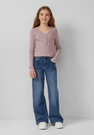 Junges Mädchen steht mit einer Hand in der Tasche, trägt ein mauvefarbenes, langärmeliges Oberteil mit Knöpfen, weite blaue Jeans und weiße Sneakers.