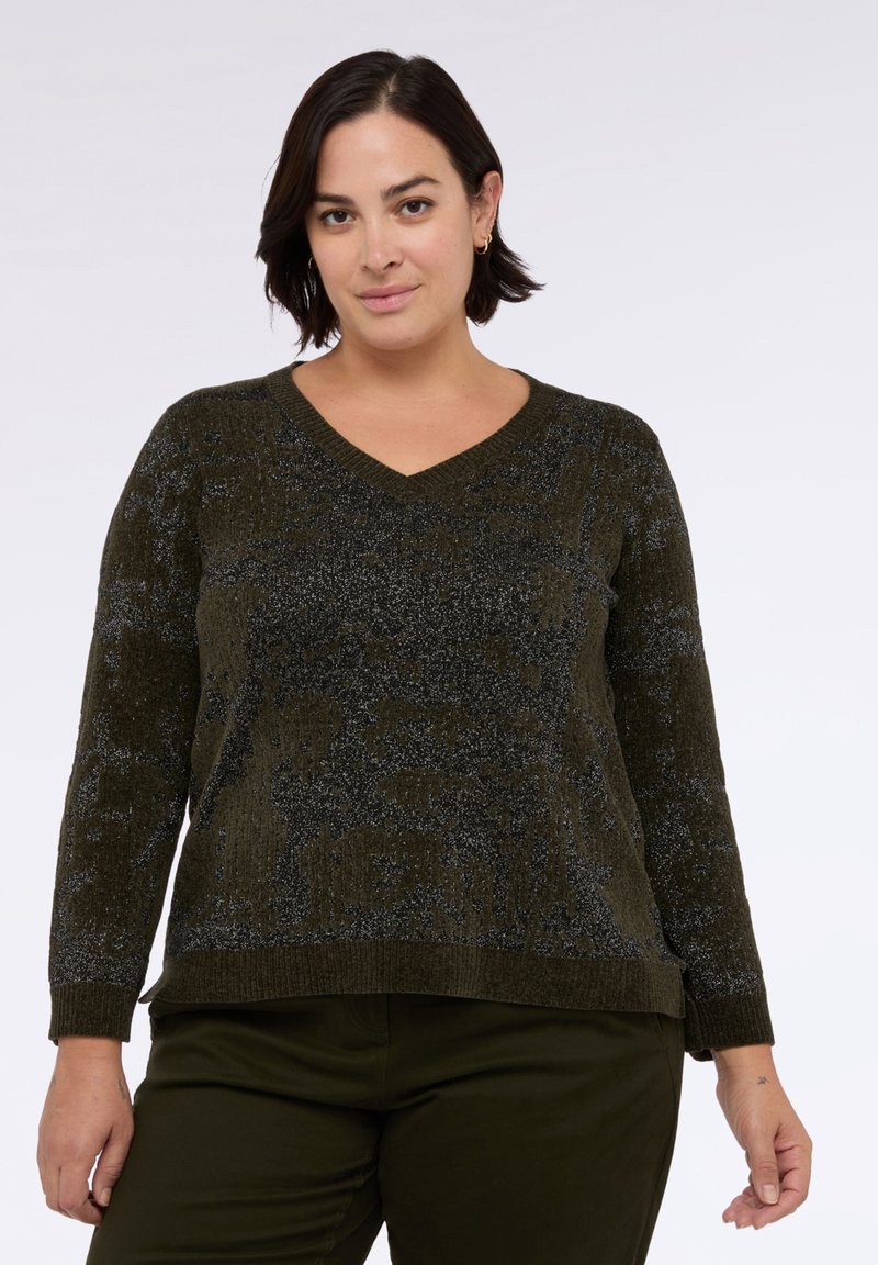 Kobieta z krótkimi ciemnymi włosami, ubrana w ciemnozielony sweter V-neck o fakturowanej powierzchni i dopasowane spodnie, stojąca na tle jasnego, jednolitego tła.