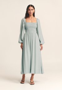 Dlhé, svetlozelené šaty so štvorcovým výstrihom, zdobeným živôtikom so záhybmi a nafúknutými dlhými rukávmi. Materiál má jemnú textúru a splývavý dizajn.