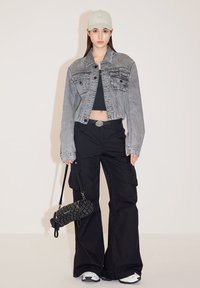 Chaqueta de mezclilla recortada en gris, camiseta sin mangas negra, pantalones cargo negros, llevando un bolso texturizado negro y usando una gorra gris claro.