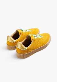 Sneakers gialli in suede con suola in gomma rigata, dettagli dorati sul tallone e lacci verdi e bianchi. Presentano un logo decorativo di lato.