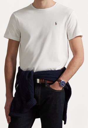 Homme portant un polo blanc à manches courtes avec un petit logo, un jean foncé, un pull bleu marine noué autour de la taille et une montre à bracelet en cuir marron.