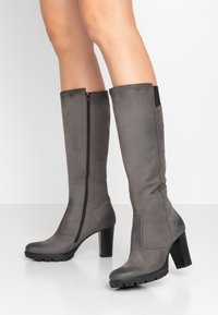 Bottes grises hauteur genou en cuir lisse, avec une fermeture éclair à l'avant, un talon épais et une semelle en caoutchouc avec un motif de bande de roulement texturé.