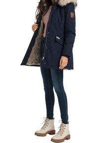 Marineparkacoat med pyntepels hætte, lynlås lukning og sidelommer. Båret over en let top og blå jeans, parret med beige snøresko.