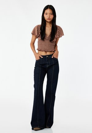 Jonge vrouw met lang zwart haar, die een bruin patroon crop top en donkerblauwe hoog getailleerde wijde spijkerbroek draagt, staand met handen in de zakken.