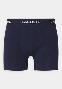 Tumši zili vīriešu bokseri ar melnu jostu, kurā ir balta "LACOSTE" apzīmējuma. Mīksts audums, pieguļoša forma un gluda tekstūra.
