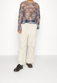 Personne portant un haut à manches longues en crochet ample bleu marine et blanc, un pantalon cargo crème et des chaussures noires à lacets, debout sur un sol blanc.