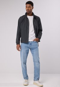 Schwarze leichte Jacke mit durchgehendem Reißverschluss, Stehkragen und elastischen Bündchen, kombiniert mit hellblauen Jeans und weißen Sneakern.