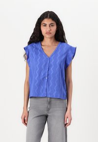 Blusa di cotone blu con scollo a V, maniche corte a volant, con un motivo a zigzag e chiusura a bottoni. Abbinata a jeans grigi.