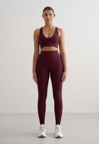 Burgundowy stanik sportowy z zamkiem błyskawicznym z przodu, w połączeniu z legginsami o wysokim stanie w tym samym kolorze. Model nosi białe sneakersy.