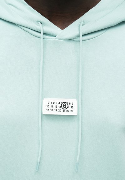 Mátově zelená mikina s kapucí a šňůrkami, s bílou štítkem nesoucím černá čísla. Vyznačuje se měkkou texturou a ležérním designem.