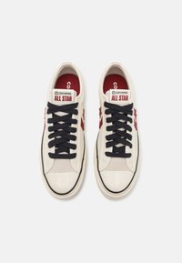 Paire de baskets blanches Converse All Star avec des lacets noirs, un logo étoile rouge sur les côtés et des bouts en caoutchouc blanc sur un fond clair.