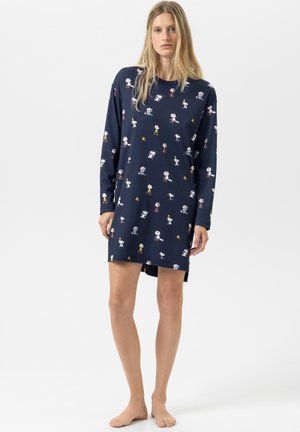 Vrouw die blootsvoets staat in een marineblauwe lange mouw jurk met Snoopy en Woodstock cartoonprints.