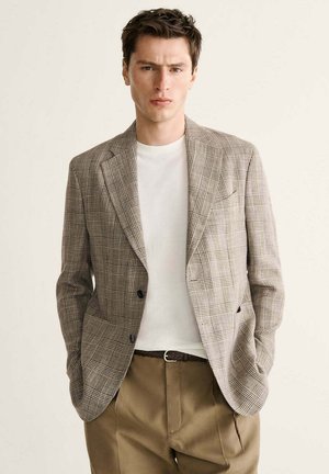 Junger Mann, der einen beigen karierten Blazer, ein weißes T-Shirt und braune Hosen trägt, steht mit den Händen in den Taschen vor einem schlichten Hintergrund.