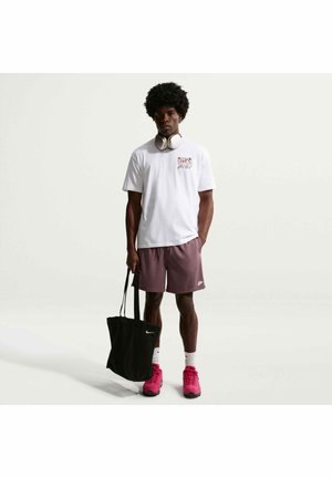 Mann trägt weißes Nike T-Shirt, bordeauxrote Shorts, pinke Schuhe, weiße Kopfhörer um den Hals, hält schwarze Nike Tragetasche, steht vor einfarbigem Hintergrund.