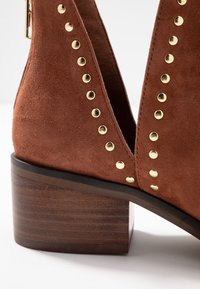Bottines en suède marron avec un large talon en bois, présentant des accents de clous dorés le long des bords et un design ouvert sur le côté.