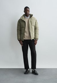 Tamsiai žalia paminkštinta striukė su aukšta apykakle, po ja dėvimas béžinis megztinis, juodos kelnės ir juodi loaferiai, išdėstyti prieš neutralų foną.