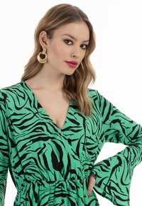 Grünes Kleid mit schwarzen Zebra-Streifen, V-Ausschnitt, elastischem Bund und Glockenärmeln. Goldene Creolen betonen den Look.