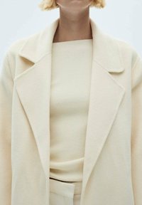 Manteau en mélange de laine crème avec un col cranté, superposé sur un top côtelé beige clair. Design minimaliste avec une texture douce.