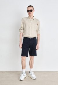 Camicia button-up in lino beige, pantaloni corti blu scuro e dettagli in bianco delle sneakers, con una tasca sulla camicia e occhiali da sole.