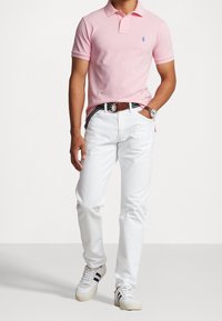 Ljusrosa polo tröja, vita jeans, randigt bälte och vita sneakers med blå accenter. Avslappnat outfit med lös passform.