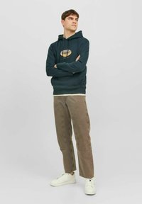 Sudadera verde oscuro con un logo en el frente, combinada con pantalones marrón claro. Zapatillas blancas completan el conjunto casual. Texturas suaves en todo.